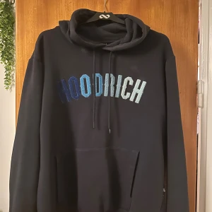 Svart hoodie från Hoodrich med blått tryck - Svart hoodie från Hoodrich i storlek M med stort broderat HOODRICH-tryck i olika blå nyanser på bröstet. Tröjan har huva med snörning och en klassisk magficka framtill. Perfekt för dig som gillar streetwear och vill ha en chill look.