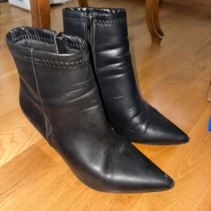 Svarta stövletter med spetsig tå - Säljer ett par svarta stövletter i skinnimitation med spetsig tå och smal klack. Skorna har dragkedja på sidan och dekorativ söm upptill. Perfekta för dig som gillar en stilren och edgy look.