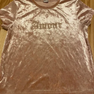 Champagnefärgad sammetstopp Lindex - Säljer en mjuk och glansig champagnefärgad topp från Lindex med korta ärmar. Toppen är i sammet och har texten 'Amour' framtill, dekorerad med små pärlor i guld. Perfekt för dig som gillar lite extra glam i vardagen.