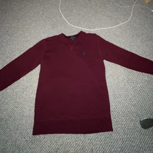 Vinröd tröja från Polo Ralph Lauren - Vinröd långärmad tröja från Polo Ralph Lauren med blå broderad logga på bröstet. Klassisk rund halsringning med snygg detaljsöm framtill. Skön passform och tillverkad i mjuk bomull, perfekt för höst och vinter.