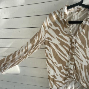Zebramönstrad skjorta från Object - Snygg skjorta från Object med beige och vita zebraränder. Modellen har lång ärm, klassisk krage och knappar framtill. Materialet är mjukt och lätt, perfekt för dig som gillar att sticka ut med coola mönster. Passformen är normal😇💕
