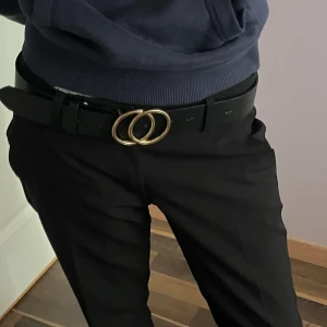 Svart bälte med guldfärgat spänne Gina Tricot - Stilrent svart bälte från Gina Tricot med ett snyggt, dubbelt guldfärgat ring-spänne. Bältet är i slätt syntetmaterial och passar perfekt till jeans eller kostymbyxor. Enkel design som lyfter vilken outfit som helst.