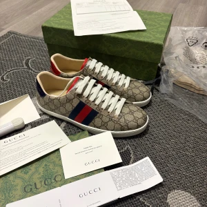 Gucci GG Supreme sneakers beige/röd/blå - Gucci sneakers i GG Supreme canvas med beige monogrammönster, vita snören och vit sula. Sidan har klassiska röd-blå ränder och hälen är i metallicrött och metallicblått. Insidan är ljusbeige och materialet är canvas med skinn-detaljer. Snygg och ikonisk modell.