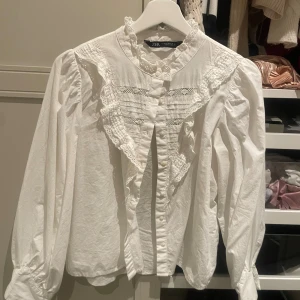 Vit blus med spets och volanger från Zara - Supersöt vit blus från Zara med romantiska volanger och spetsdetaljer längs bröstet och kragen. Långärmad modell med puffiga ärmar och knappar framtill. Perfekt för dig som gillar en feminin och trendig stil.