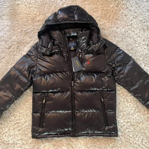 Svart pufferjacka från Polo Ralph Lauren - Svart pufferjacka från Polo Ralph Lauren med glansig finish, huva och två dragkedjefickor. Klassisk röd Polo-logga på bröstet och snygga detaljer vid kragen. Jackan har lång ärm och är perfekt för kalla dagar.