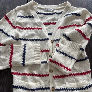 Randig pippi kofta från Rut m.fl.  - Snygg stickad kofta i offwhite med röda och marinblå ränder från Rut m.fl. Koftan har bruna armbågslappar, två fickor fram och knäpps med knappar. Perfekt för lager-på-lager och chill stil. 