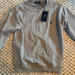 Grå sweatshirt från Polo Ralph Lauren - Snygg grå sweatshirt från Polo Ralph Lauren med klassisk rund halsringning och broderad logga på bröstet. Tröjan har ribbade muddar vid ärmslut och nederkant. Perfekt för en chill och stilren look. I storlek M Men sitter mer som s