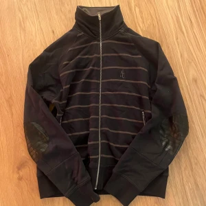 Svart randig jacka från Emilio - Svart jacka från Emilio med diskreta horisontella ränder och broderad logga på bröstet. Jackan har hel dragkedja, hög krage och förstärkta detaljer i skinnimitation på armbågarna. Perfekt för dig som gillar stilren och sportig look.