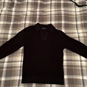 Pikétröja  - Svart stickad pikétröja från Riley med klassisk krage och v-ringad hals. Tröjan har lång ärm och en stilren, enkel design som passar perfekt till jeans. 