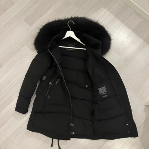  Cedrico jacka - Säljer en svart dunjacka från Cedrico med fluffig päls på huvan. Jackan har flera dragkedjor, knappar och snörning nedtill. Den är lång, har en snygg quiltad design och passar perfekt när det är kallt ute. Huvan är stor och ger extra värme och stil.