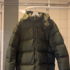 Grön pufferjacka Polo Ralph Lauren XL - Mörkgrön pufferjacka från Polo Ralph Lauren i storlek XL. Jackan har huva, dragkedja framtill och en bröstficka med dragkedja. Klassisk orange Polo-logga på bröstet. Perfekt för kyla