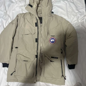 Beige dunjacka från Canada Goose L - Säljer en beige Canada Goose dunjacka i storlek L. Jackan har flera fickor med lock, huva och klassisk Canada Goose patch på bröstet. Quiltad insida och dragkedja framtill. Perfekt för kalla vinterdagar och riktigt snygg streetstil.