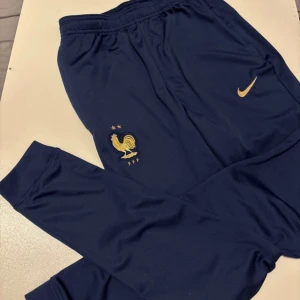 Blå Nike FFF träningsbyxor slim fit - Mörkblå träningsbyxor från Nike med FFF-logga och guldiga detaljer. Slim fit-modell i Dri-Fit polyester, perfekt för fotboll eller träning. Elastisk midja och broderade loggor på båda sidorna ger en sportig och clean look.