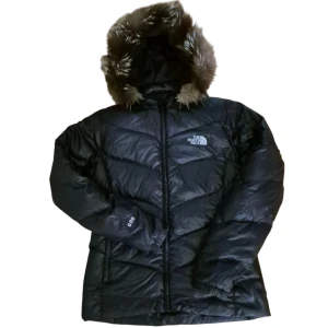 Svart dunjacka från The North Face  - Säljer en svart dunjacka från The North Face med huva och fuskpäls. Jackan har dragkedja framtill, quiltad design och logga på bröstet. Perfekt för kalla dagar med sin fluffiga och varma känsla. Klassisk modell med lång ärm och snygg passform.