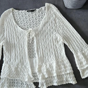 Vit spetsig kofta från KappAhl - Superfin vit kofta från KappAhl med spetsdetaljer och knyt framtill. Koftan har 3/4 lång ärm med volang vid ärmslut och nederkant, samt ett genomgående hålmönster som ger en romantisk vibe. Perfekt för dig som gillar boho-stil och vill ha något luftigt och snyggt.