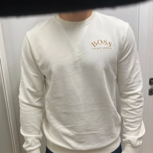Vit sweatshirt från Hugo Boss - Vit sweatshirt från Hugo Boss med broderad logga på bröstet och snygga guldiga detaljer längs ärmarna. Tröjan har rund halsringning, ribbade muddar och är tillverkad i mjuk bomull. Perfekt för en clean och stilren look.