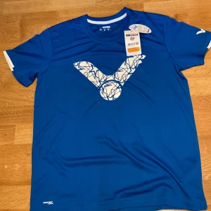Victor - Blå sportig t-shirt - Badminton  - Blå t-shirt från Victor med stort vitt grafiskt tryck på bröstet. Perfekt för träning eller sport, tillverkad i lätt och snabbtorkande polyester. Klassisk passform med korta ärmar och diskreta vita detaljer vid ärmslut.