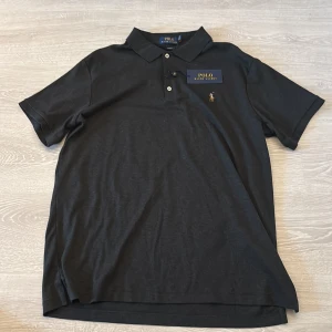 Svart piké från Polo Ralph Lauren XL - Svart pikétröja från Polo Ralph Lauren i slim fit-modell. Klassisk krage, korta ärmar och knappar framtill. Ikonisk broderad logga med polospelare på bröstet. Mjuk bomullskvalitet som känns skön mot huden. Perfekt för dig som gillar stilren och enkel design.