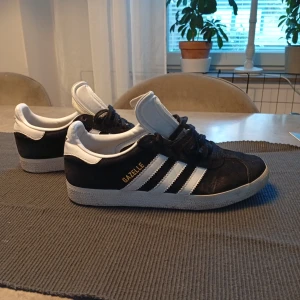 Adidas Gazelle svart/vit mocka - Säljer ett par klassiska Adidas Gazelle sneakers i svart mocka med vita detaljer och de ikoniska tre ränderna på sidan. Skorna har snörning, vit sula och guldtexten 'GAZELLE' på sidan. Perfekta för dig som gillar retrostil och streetwear.
