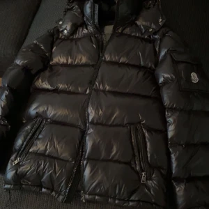 Svart Moncler pufferjacka med huva - Säljer en svart pufferjacka från Moncler med glansig finish, huva och flera dragkedjefickor. Jackan har en klassisk Moncler-logga på ärmen och quiltad design. Perfekt för kalla dagar och riktigt snygg till streetwear-stilen.