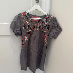 Broderad grå blus från Bondelid - Grå blus från Bondelid med färgglada broderier i rött, gult och svart framtill och på ryggen. Modellen har korta puffärmar med knappdetalj och rund halsringning. Tillverkad i mjuk bomull, perfekt för dig som gillar boho-vibbar och snygga detaljer. 