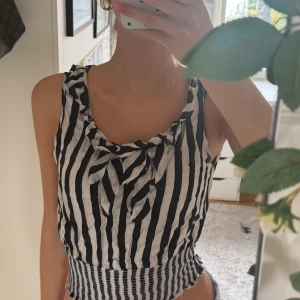 Randig svartvit linnetopp med rosett - Säljer en snygg ärmlös linnetopp med svarta och vita vertikala ränder. Toppen har en söt rosett vid halsringningen och smockad, vågig nederkant som ger en cool och trendig look. Perfekt för dig som gillar att sticka ut med mönster och detaljer.