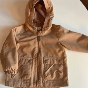 Beige vindjacka från Newbie med huva - Säljer en beige vindjacka från Newbie i storlek 80. Jackan har huva, dragkedja framtill och två stora fickor med knappar. Perfekt för blåsiga dagar och enkel att matcha med andra plagg.