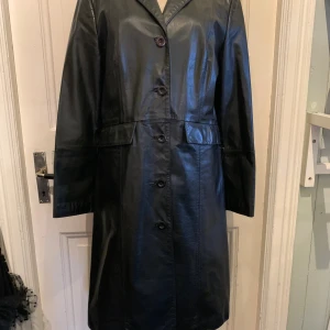 Svart skinnrock från Sisley, st L - Säljer en svart lång skinnrock från Sisley i äkta läder. Riktig Vera Pelle. Rocken har klassisk krage, knäppning med fyra knappar framtill och två fickor med lock. Snygg, tidlös modell som ger en cool vibe till din outfit.