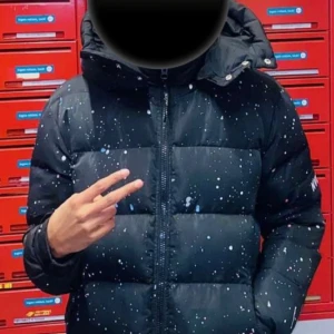 Svart pufferjacka från NVLTY med färgstänk - Svart pufferjacka från NVLTY med coola färgstänk i vitt, rosa och blått. Jackan har huva med tryckknappar, dragkedja framtill och fickor med dragkedja. Perfekt för dig som vill sticka ut med en unik streetstyle-look.