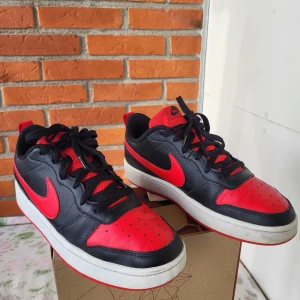 Nike Court Borough Low 2 sneakers svart/röd strl 40 - Snygga Nike sneakers i svart och rött med klassisk swoosh på sidorna. Skorna har lågt skaft, snörning och vit mellansula med röd yttersula. Ovandelen är i skinn och textil, med ventilerande hål på tån. Perfekta för dig som gillar sportig och street-inspirerad stil.