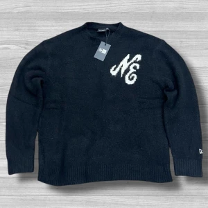 Stickad tröja från New Era - Svart stickad tröja från New Era med vit NE-logga på bröstet och broderad logga på ärmen. Klassisk rund halsringning och ribbade muddar. Perfekt för dig som gillar streetwear och vill ha en stilren look. Storlek M men passar L