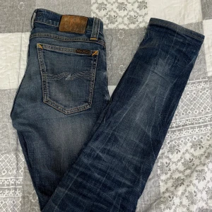 Blå jeans från Nudie Jeansco, 26/34 - Snygga blå jeans från Nudie Jeansco i storlek 26/34. Klassisk femficksmodell med orange kontrastsömmar och coolt vågmönster på bakfickan. Jeansen har en rak passform och är tillverkade i slitstarkt denimtyg. Perfekt för dig som gillar en tidlös och avslappnad stil.