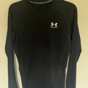 Svart långärmad träningströja UA M - Svart långärmad träningströja från Under Armour med HeatGear-teknologi. Tight passform och diskret logga på bröstet och ryggen. Perfekt för intensiva träningspass och sportaktiviteter. Materialet är syntet och designen är stilren och sportig.