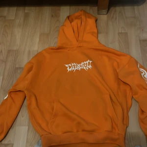Orange hoodie med draktryck från FSBN - Säljer en orange hoodie från FSBN med fet drake och coola tribal-tryck i vitt på ryggen och ärmarna. Hoodien har huva, magficka och är gjord i mjuk bomullsmix. Perfekt för dig som vill sticka ut med streetstyle vibes.