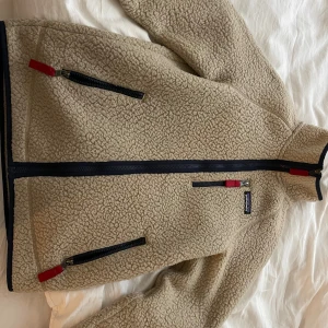 Beige teddyjacka från Patagonia - Säljer en beige teddyjacka från Patagonia med hög krage och svarta detaljer. Jackan har flera dragkedjeförsedda fickor med röda zip-pullers och ett snyggt Patagonia-märke på bröstet. 