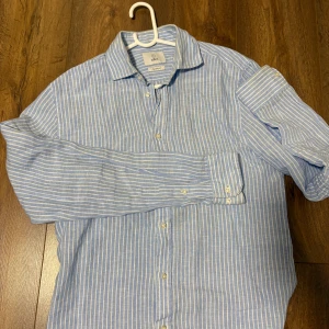 Blåvit randig linneskjorta från Bläck - Snygg blå och vit randig skjorta från Bläck i linnematerial. Skjortan har klassisk krage, långa ärmar och knäppning framtill. Perfekt för en fräsch och avslappnad stil under varmare dagar. Några fläckar på inre kragen vilket jag kan försöka få bort ifall att.