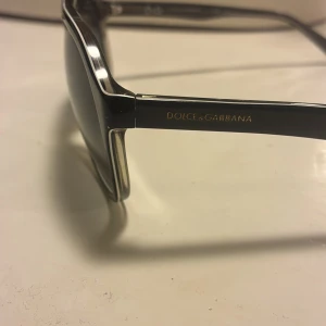 Svarta solglasögon från Dolce & Gabbana - Snygga svarta solglasögon från Dolce & Gabbana med klassisk pilotform och diskreta silvriga detaljer. Bågarna är i blank plast och har märkets logga i guld på sidorna. Perfekta för att lyfta din stil med en lyxig touch.