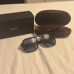 Svarta Tom Ford glasögon - Snygga svarta glasögon från Tom Ford med fyrkantig form och mörka glas. Ramen är svart med guldiga T-detaljer på sidorna. Kommer med originalfodral och box. Perfekt accessoar för att lyfta din stil.