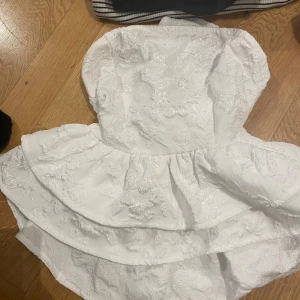 Vit axelbandslös kortklänning med struktur - Superfin vit axelbandslös kortklänning med volanger och broderade blommor i strukturerat tyg. Klänningen har en markerad midja och en söt omlottknytning i ryggen. Perfekt för sommarens alla tillfällen!