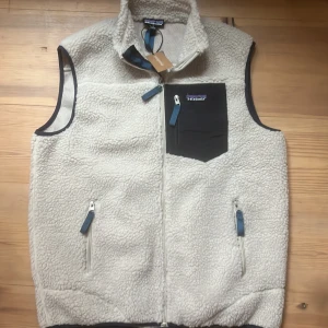 Beige fleeceväst från Patagonia - Säljer en beige fleeceväst från Patagonia med hög krage och dragkedja framtill. Västen har en svart bröstficka med dragkedja och två sidofickor med blå detaljer. Perfekt för lager-på-lager och outdoor vibes. Ny!