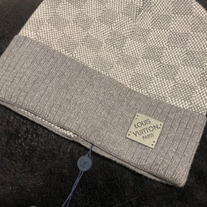 Grå rutig mössa från Louis Vuitton - Snygg grå mössa från Louis Vuitton med klassiskt rutigt mönster och ribbad kant. Märkespatch framtill och diskret LV-tag inuti. Tillverkad i mjukt material som känns lyxigt och passar perfekt för kalla dagar.