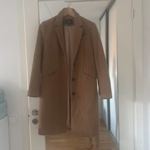 Beige kappa från GANT XS - Snygg beige kappa från GANT i storlek XS. Klassisk rak modell med krage, två fickor och knäppning framtill. Materialet är 75% ull, 19% polyamid och 6% kashmir, vilket gör den både mjuk och varm. Perfekt för dig som gillar stilrena och tidlösa plagg.