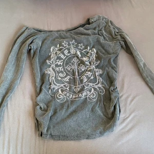 Grå offshoulder topp från Romwe - Säljer en grå långärmad offshoulder topp från Romwe med ett stort tryck framtill i vitt med stjärnor, kors och text. Toppen är ribbad och har en tight passform som ger en fint vibe. Perfekt för dig som gillar ha det lowkey och glittrig 