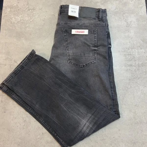Svart-Grå straight jeans från SMOG - Säljer ett par svart-grå jeans från SMOG i straight fit. Jeansen har klassisk femficksdesign, normal midja och snygga slitningar för en cool look. Materialet är jeans och färgen är mörkgrå med ljusare tvättade partier. Perfekta för dig som gillar en avslappnad stil.
