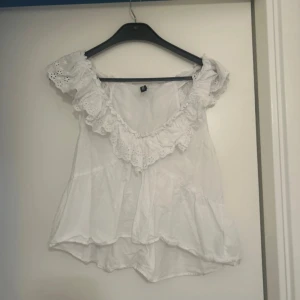 Vit blus med volang från H&M Divided - Supersöt vit blus från H&M Divided i storlek S. Blusen har offshoulder-design med breda volanger och broderade spetsdetaljer längs halsringningen. Tillverkad i mjuk bomull, perfekt för sommarens alla tillfällen. Fluffig och luftig passform med romantisk vibe.