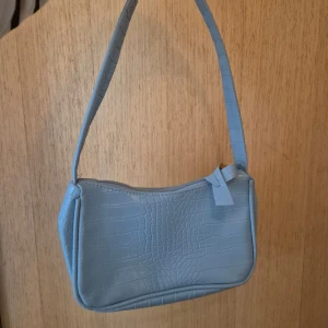 Ljusblå axelväska i krokopräglat skinn - Snygg ljusblå axelväska med croco-mönstrad yta och dragkedja upptill. Väskan har en stilren, avlång form och bred axelrem. Perfekt för dig som vill ha en trendig accessoar med cool struktur. Märke ej angivet.