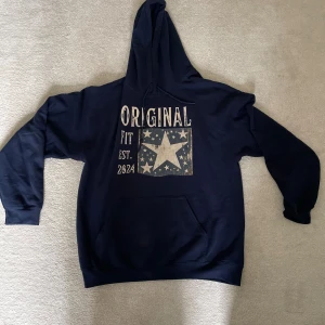 Mörkblå hoodie - Mörkblå hoodie från Original Fit med stor vit stjärna och texten 'Original Fit Est. 2024' framtill. Tröjan har huva med snörning och en stor magficka. Jätte fin hoodie! Knappt använd 