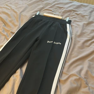 Palm Angels svarta mjukisbyxor med vita ränder - Svarta mjukisbyxor från Palm Angels med vita sidoremsor och broderad logga på vänster ben. Byxorna har elastisk midja och raka ben, tillverkade i polyester för en sportig vibe. Perfekta för en chill och trendig streetwear-look. Dom är köpte här på Plick så kan inte garantera äkthet därför det låga priset 