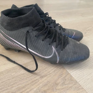 Svarta Nike fotbollsskor med snörning - Säljer ett par svarta Nike fotbollsskor med högt skaft och snörning. Skorna har en diskret silverfärgad swoosh och är tillverkade i syntetmaterial med texturerad yta för bättre bollkontroll. Perfekta för dig som vill ha stilrena och funktionella skor på planen.