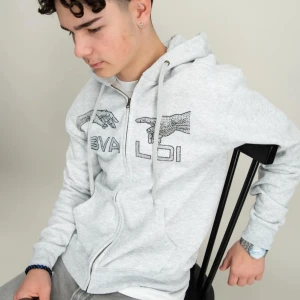 Grå hoodie från Bavaldi med dragkedja - Snygg ljusgrå hoodie från Bavaldi med dragkedja och huva. Tröjan har coolt tryck med händer och text framtill, samt snörning i huvan. Perfekt för chill dagar eller när du vill ha en avslappnad stil. Den är lite söndrig i ena handen som man ser på bild 2 annars är det bra skick. Pris kan diskuteras!!